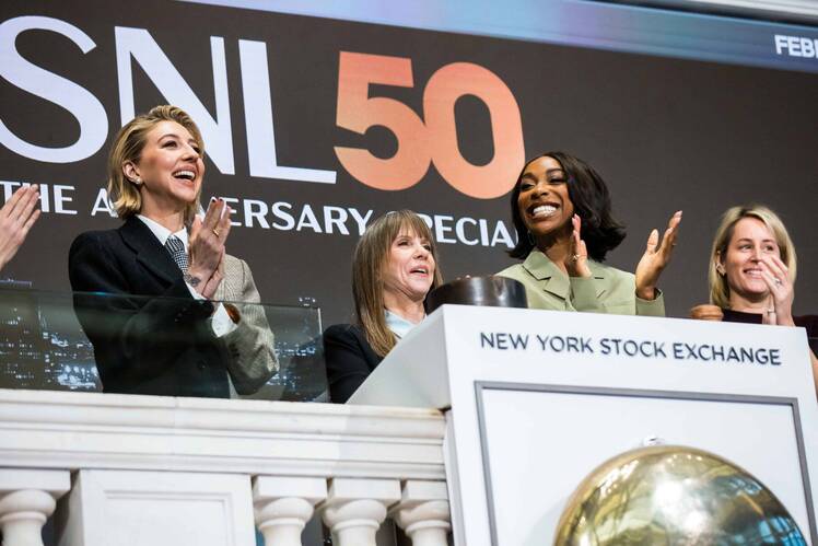 Heidi Gardner claps alongside fellow cast members at the New York Stock Exchange during SNL’s 50th anniversary celebration, standing at the podium with the event’s logo visible behind them.