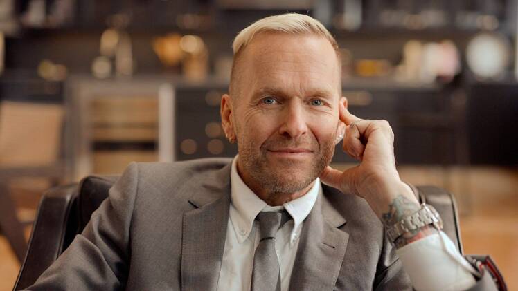 Bob Harper sits in a chair wearing a gray suit and tie, resting his chin on his hand with a slight smile
